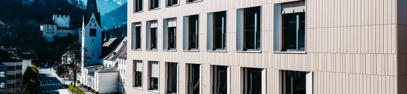 Modern building with light facade and many windows overlooking a street, church tower, castle ruin and mountains in Feldkirch (Bärenhaus).