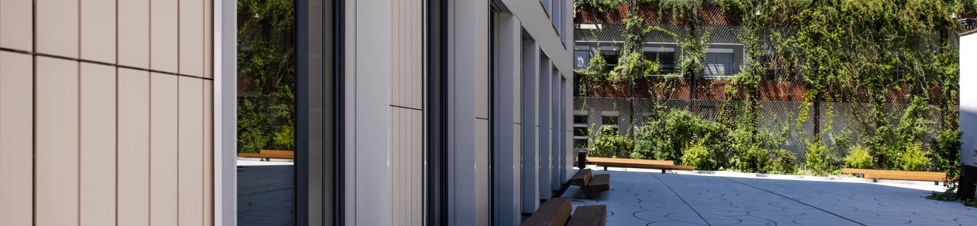 Modernes Gebäude mit hellen Fassaden und großen Fenstern, gegenüber ein begrüntes Gebäude, davor eine Terrasse mit Holzbänken (Baerahus, Feldkirch).